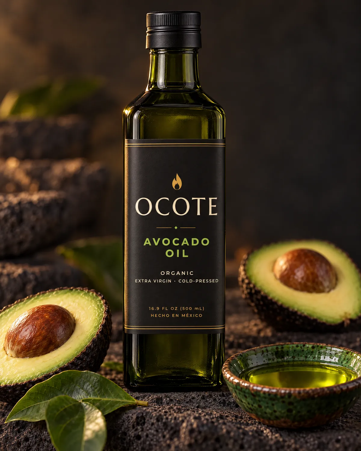 A tall square bottle of OCOTE organic extra virgin avocado oil beside halved Hass avocados and a green clay dish of avocado oil.